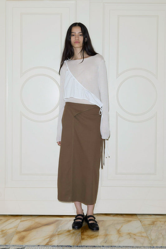 A woman with long dark hair stands indoors, wearing a beige top layered with an asymmetrical white wrap and the Deiji Studios Crossover Skirt - Carob, paired with black sandals, posing before cream paneled doors.