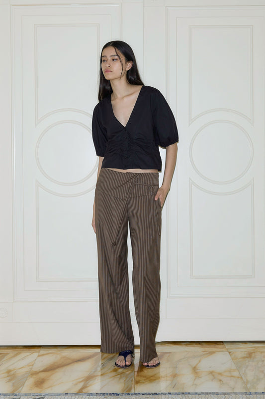 A woman with long dark hair stands indoors on a marble floor in front of white paneled doors, wearing the Folded Wool Pant—brown pinstriped wide-leg trousers—paired with a black V-neck top and dark sandals.