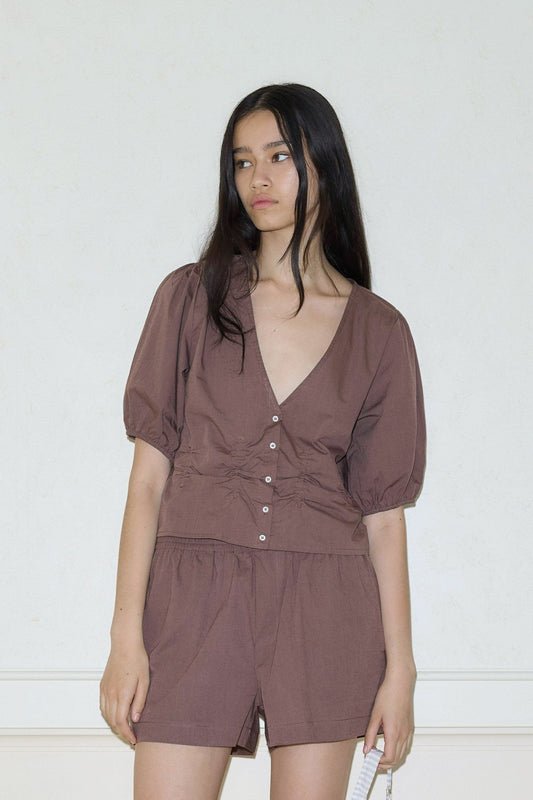 A woman with long dark hair wears the Draw Top, a brown short-sleeved, buttoned blouse with a deep V-neck, paired with matching shorts. She stands against a plain light wall, holding a white object in her left hand.