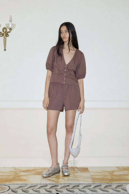 A woman stands indoors by a light wall, wearing an Organic Cotton Boxer in brown, silver shoes, and holding a small silver handbag. A wall sconce appears on the left.