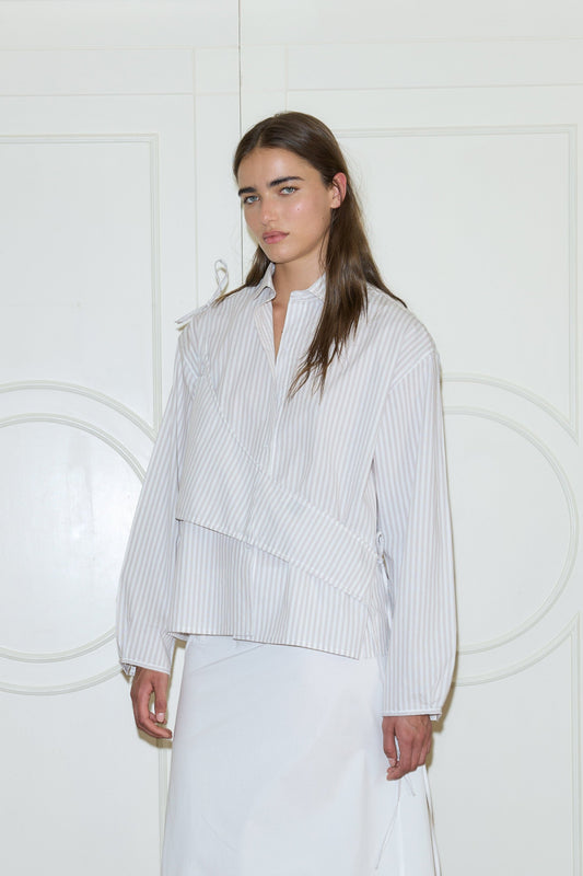 A woman with long brown hair stands in front of white paneled doors, wearing Deiji Studios' Cotton Sash - Lake Stripe blouse with a diagonal stripe design and a plain white skirt. She has a neutral expression.