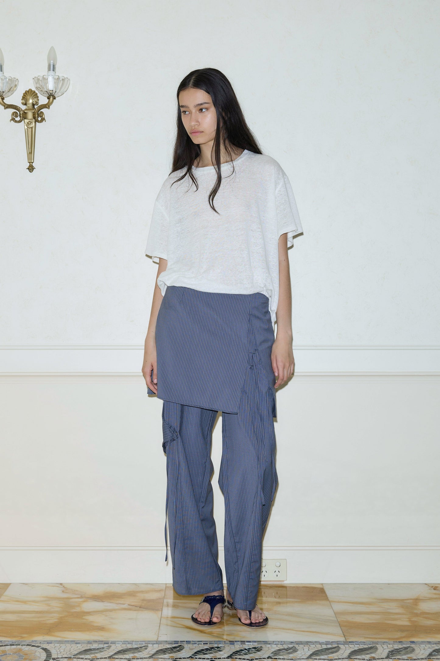 A woman with long dark hair stands indoors by a white wall, wearing a loose white T-shirt, the Two Ties Skirt - Navy Stripe, and black sandals. A brass wall sconce is visible to her left.