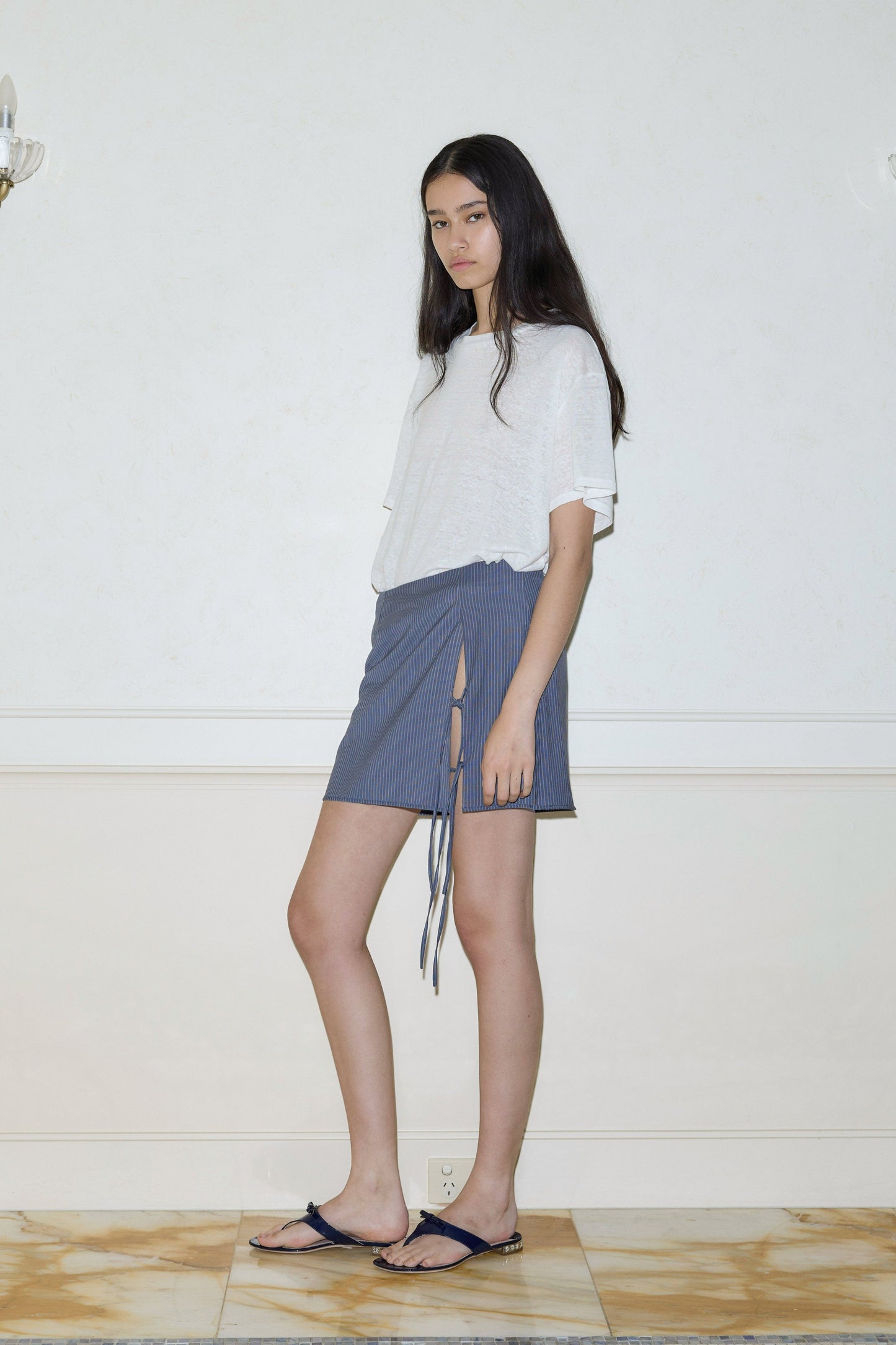 A young woman stands indoors on a marble floor, calmly facing the camera. She wears a loose white top, dark sandals, and the Two Ties Skirt - Navy Stripe: a blue wrap skirt with stripes, front tie, and slit.