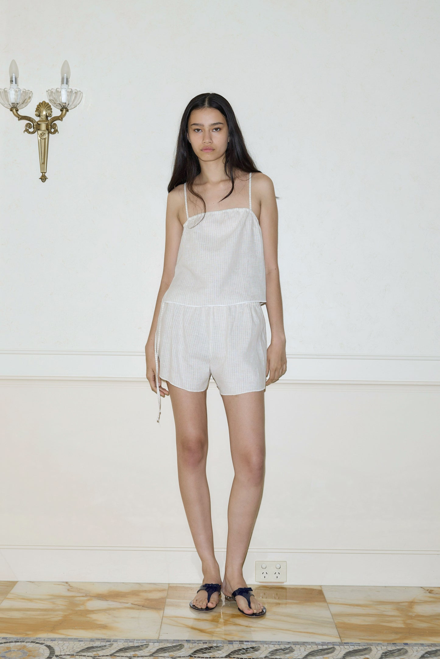 A woman with long dark hair stands indoors against a pale wall, wearing the Babylock Linen Top - Linen Story Stripe from Deiji Studios with matching shorts. She has on black sandals and stands on a marble floor near a wall sconce.
