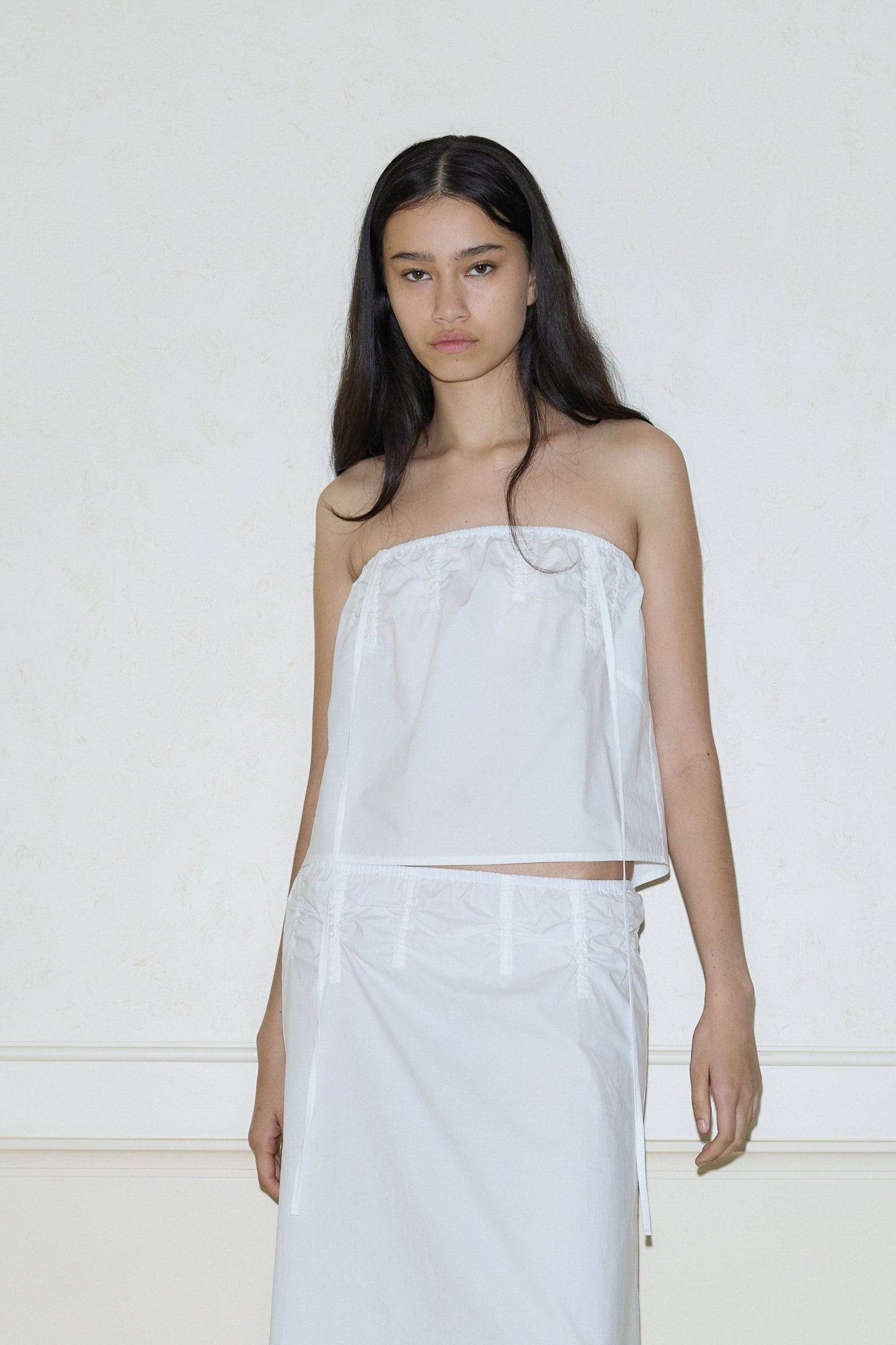A woman with long dark hair wears the Deiji Studios Tube Top - White and matching skirt, standing against a light-colored wall and looking at the camera with a neutral expression.