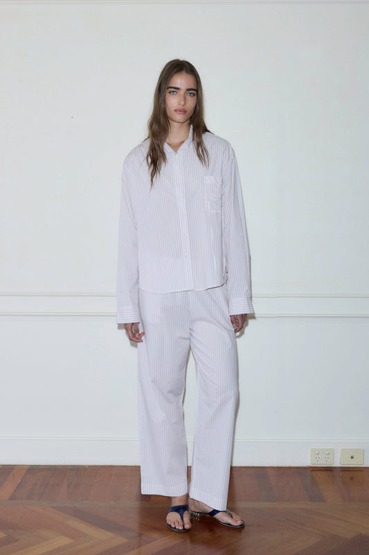 A person with long hair stands indoors against a plain white wall and wooden floor, wearing the Pocket Set—loose, light-colored, striped pajamas—and dark sandals.