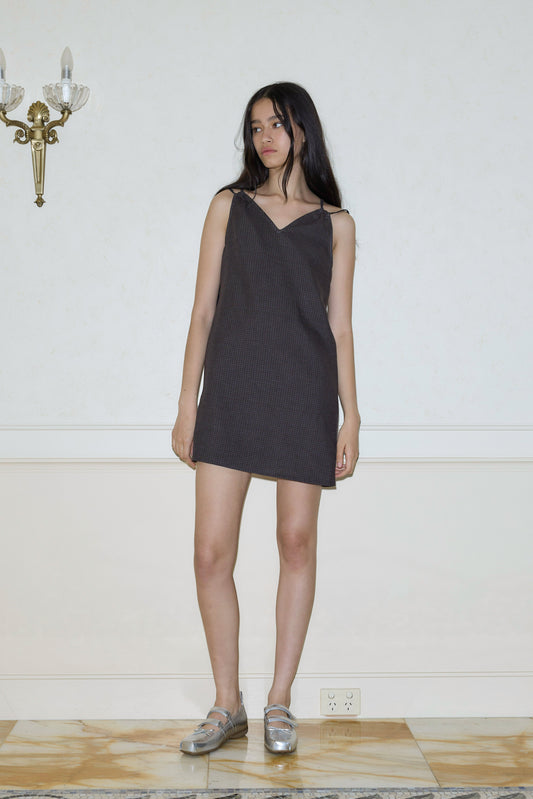 A woman with long dark hair wears the sleeveless Looped Dress - Slate Check and silver shoes, standing indoors against a white wall with a gold wall sconce and marble flooring.
