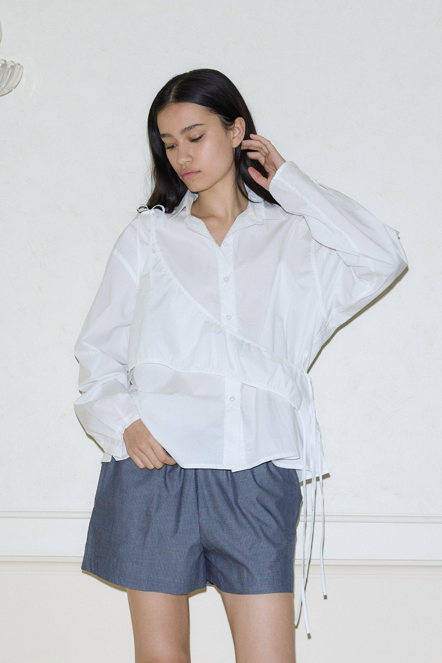 A woman with long dark hair stands against a light wall, wearing the Deiji Studios day shirt—a loose white button-up with drawstring details—and dark gray shorts, looking down with a neutral expression.