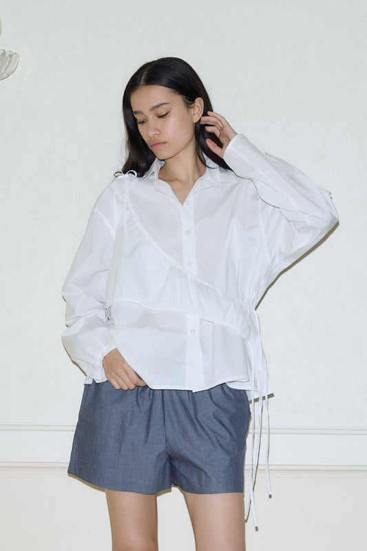 A woman stands by a light wall wearing the day shirt—a loose white button-up with drawstring details—and blue-gray shorts. She looks down, one hand on her head and the other in her pocket.