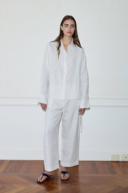 A woman with long brown hair stands barefoot indoors on a hardwood floor, wearing the Tie Split Set—a loose-fitting white button-up shirt and matching wide-leg pants—against a light-colored wall.