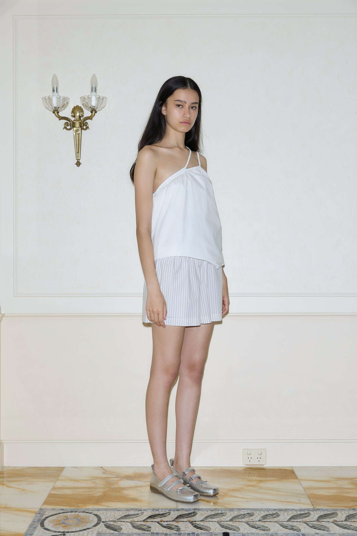 A woman with long dark hair stands indoors by a white wall under a decorative sconce, wearing a white asymmetrical top and Organic Cotton Boxer - Lake Stripe shorts with an elastic waist, silver shoes, on marble flooring and a patterned rug.