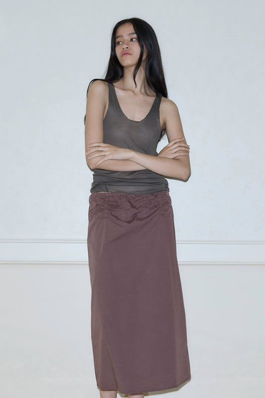A woman with long dark hair stands against a light wall, arms crossed, wearing a sleeveless brown top and the Channel Skirt—a long, textured brown skirt. She looks slightly to the side with a neutral expression.