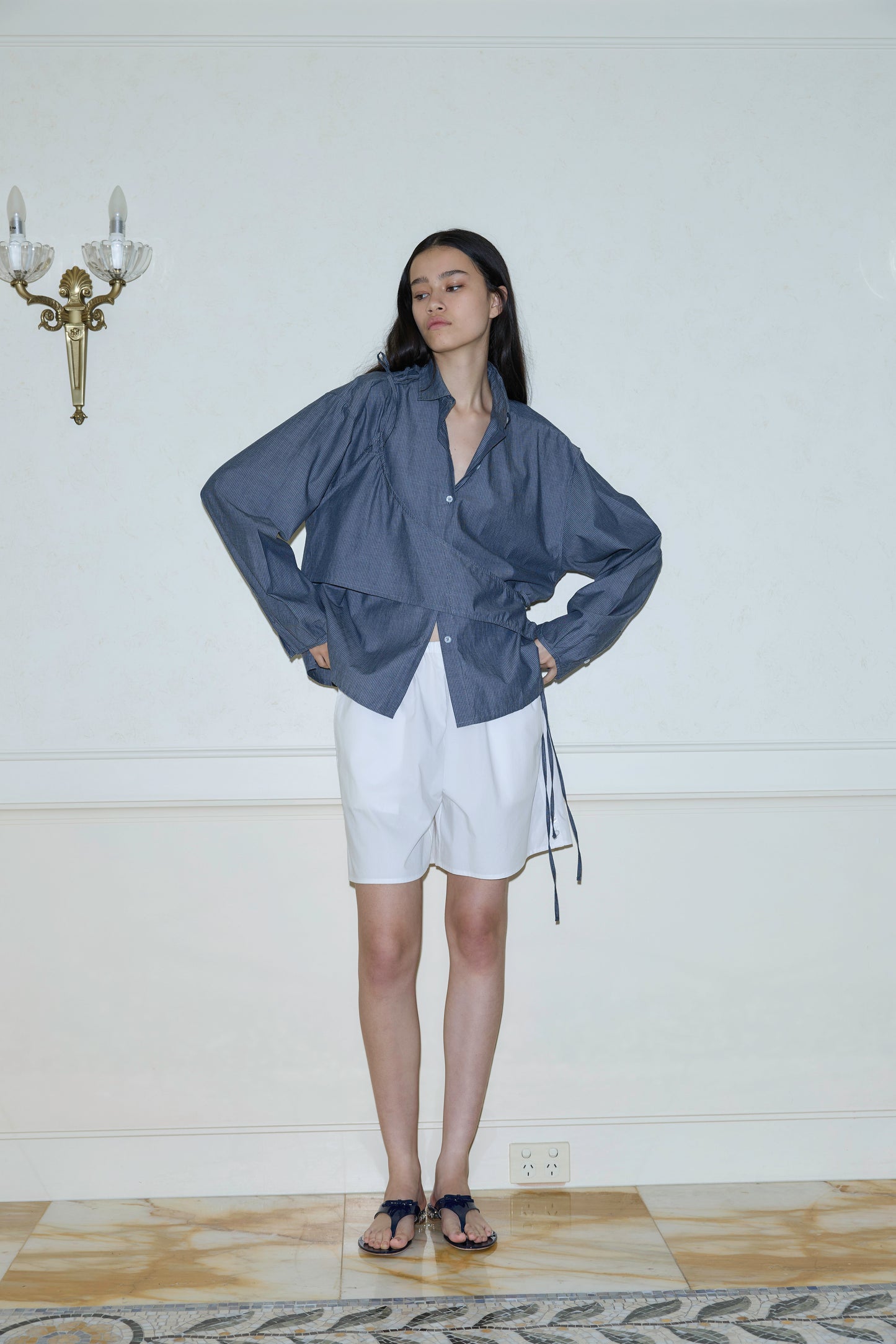 A woman with long dark hair stands indoors against a white wall, wearing Button Shorts in white, a blue long-sleeve wrap blouse, and dark sandals. Her hands are on her hips, and a wall sconce appears to her left.