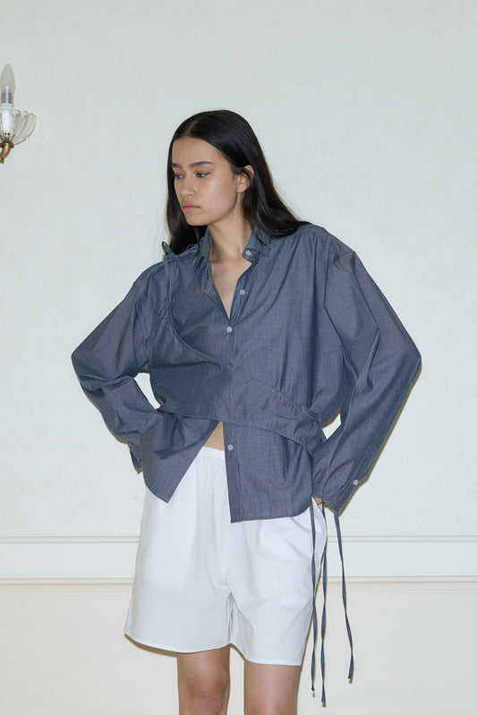 A woman stands indoors against a light wall, wearing a loose-fitting blue pinstripe Cotton Sash shirt with long sleeves and white shorts. Her hands rest on her hips as she gazes to the side with a neutral expression.