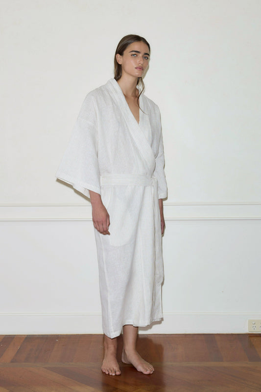 A barefoot person stands indoors on a wooden floor, dressed in the 02 Robe—a long, light-colored garment with loose sleeves—against a plain white wall, gazing slightly to the side.
