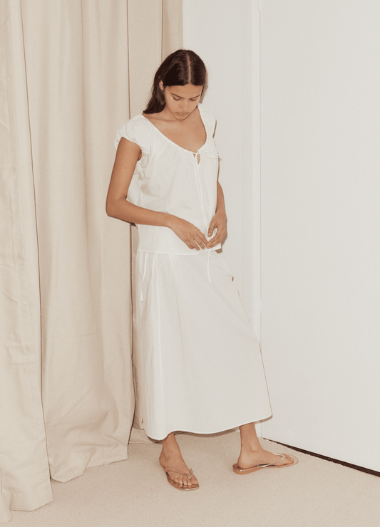 A woman stands indoors barefoot on a light carpet near beige curtains, wearing a loose-fitting white top and the Pintuck Skirt. She looks down with her hands resting near her waist against a white wall.