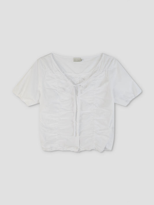 The Crinkle Top - White is a short-sleeved, white button-up top featuring a ruched front, tie detail at the neckline, and a slightly cropped hem, displayed against a plain light background.