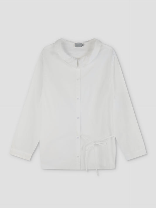 The Left Shirt - White is a lightweight, long-sleeve button-up with a rounded collar, relaxed fit, and an adjustable bow side tie at the waist, shown flat on a light background.