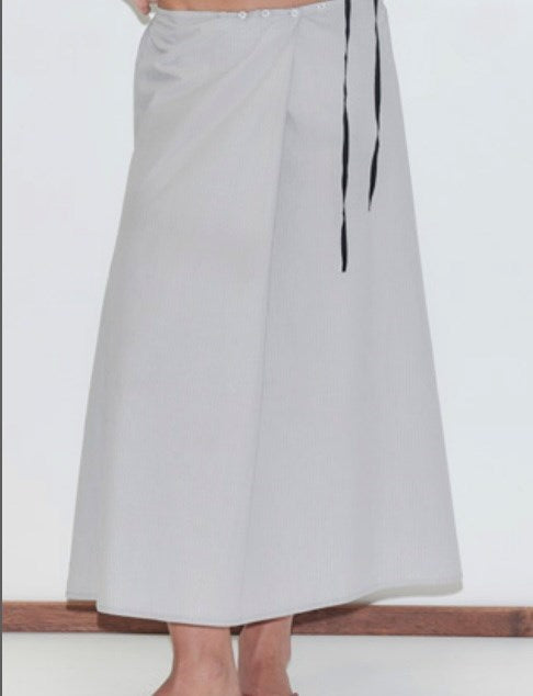 A person is modeling the Wrap Skirt, a long, light gray skirt featuring a black waist tie, while standing barefoot against a plain white background.