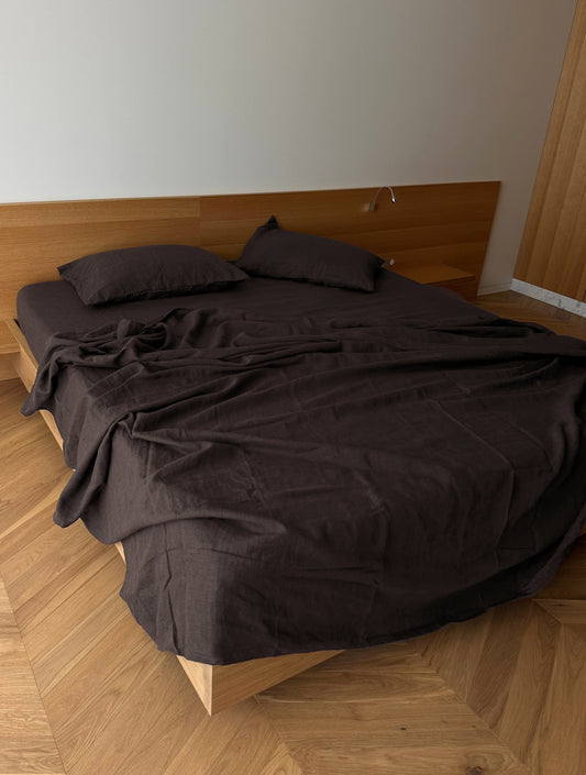 A minimalist bedroom features a modern bed with Deiji Studios’ hypo-allergenic Pillow slip set - Bark, brown sheets, a wooden headboard, light wood flooring, and plain white walls. The linen bedding looks slightly rumpled.