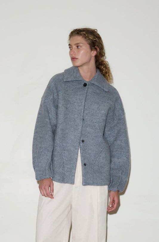 A person with wavy, light brown hair wears the Boiled Wool Coat in Blue Grey and cream-colored pants, standing against a plain white background and looking slightly to the side.