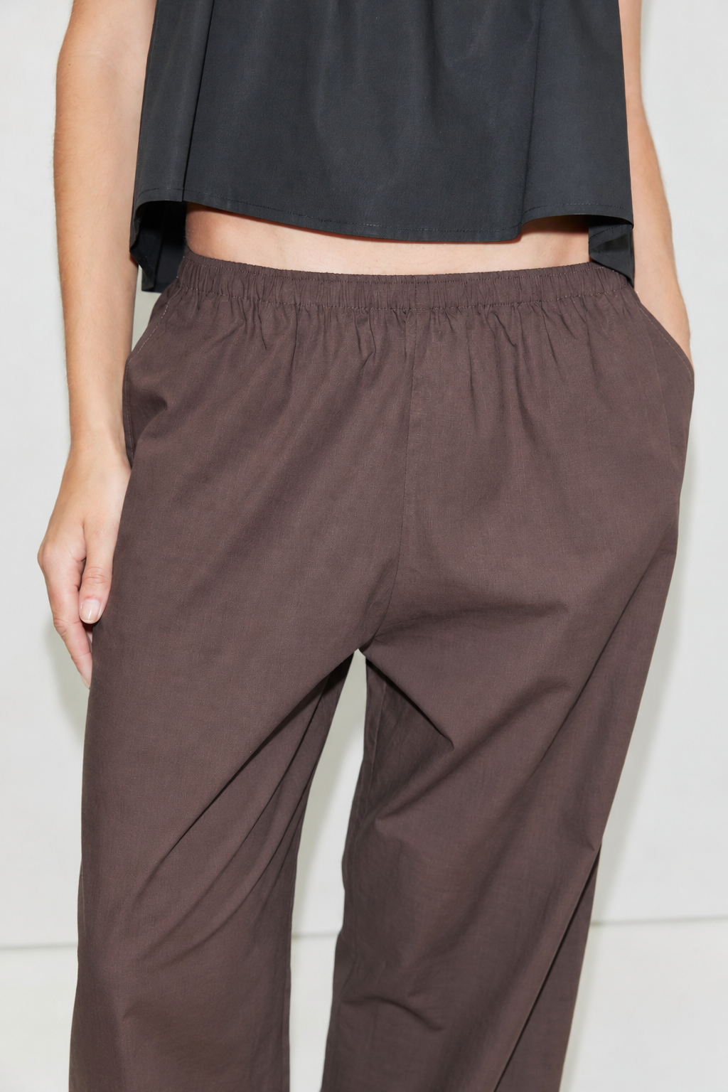 A person wearing the Ease Trouser - Toast by Deiji Studios, ankle length with a loose fit and elastic waistband, paired with a black top, shown from the torso down with one hand in their pocket.