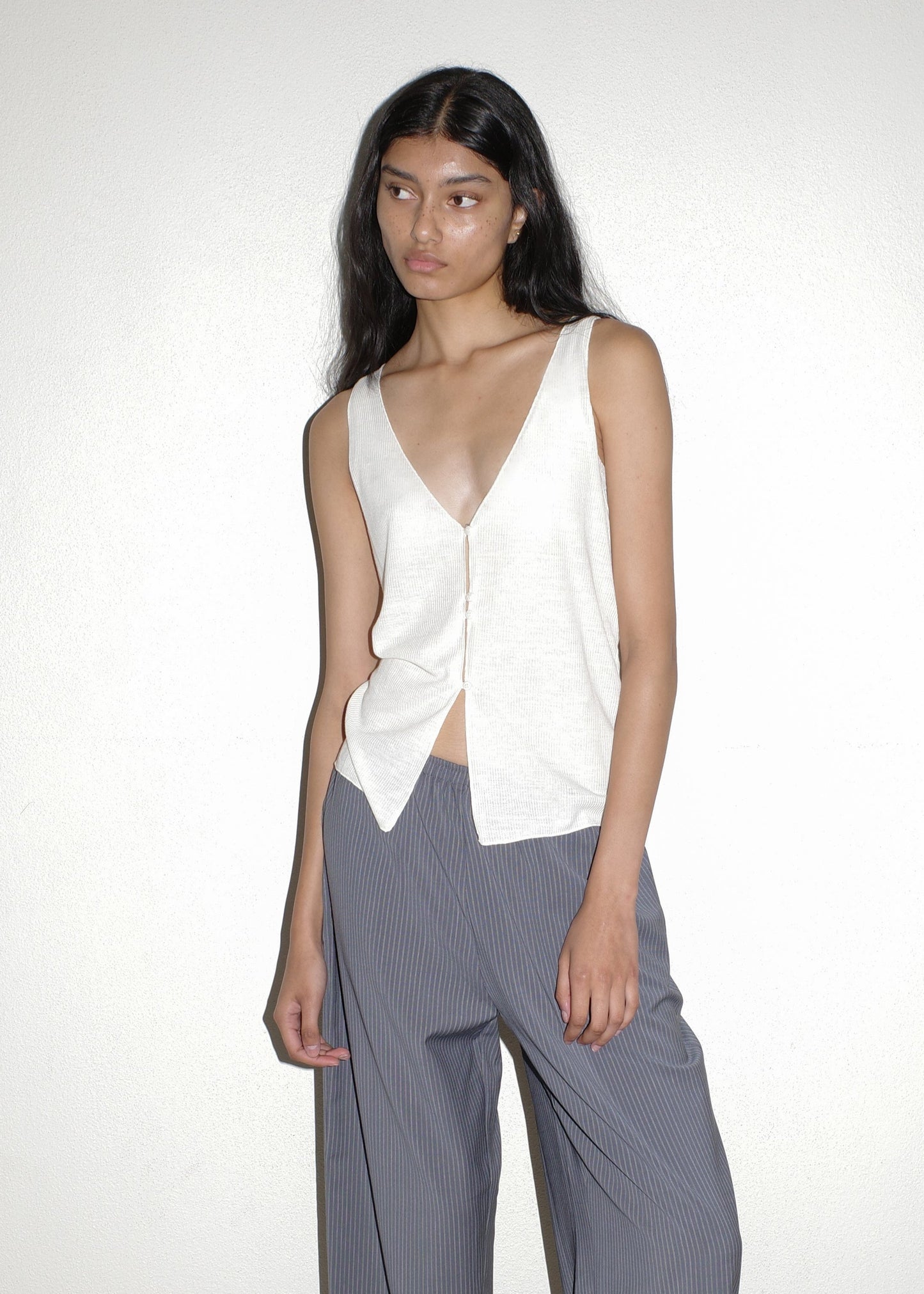 A person with long dark hair wears the Button Up Knit Tank - Off White and loose-fitting gray pinstripe pants, standing against a plain light-colored wall.