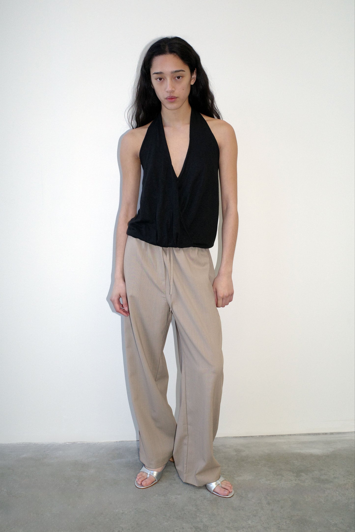 A person with long dark hair stands against a white wall, wearing a sleeveless black halter top and the Low Pocket Pure Wool Pant - Dove Stripe, paired with silver sandals. They have a neutral expression and relaxed posture.