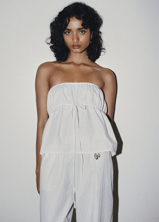 A person with curly dark hair stands against a plain wall, wearing the Strapless Cotton Top—white and light beige pinstriped, loose-fitting, with a drawstring waist—paired with matching pants.