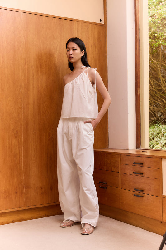 A woman stands indoors by a wood-paneled wall, wearing The Multi Strap Top - White from Deiji Studios and matching wide-legged pants, her hand in her pocket as she looks away from the camera.