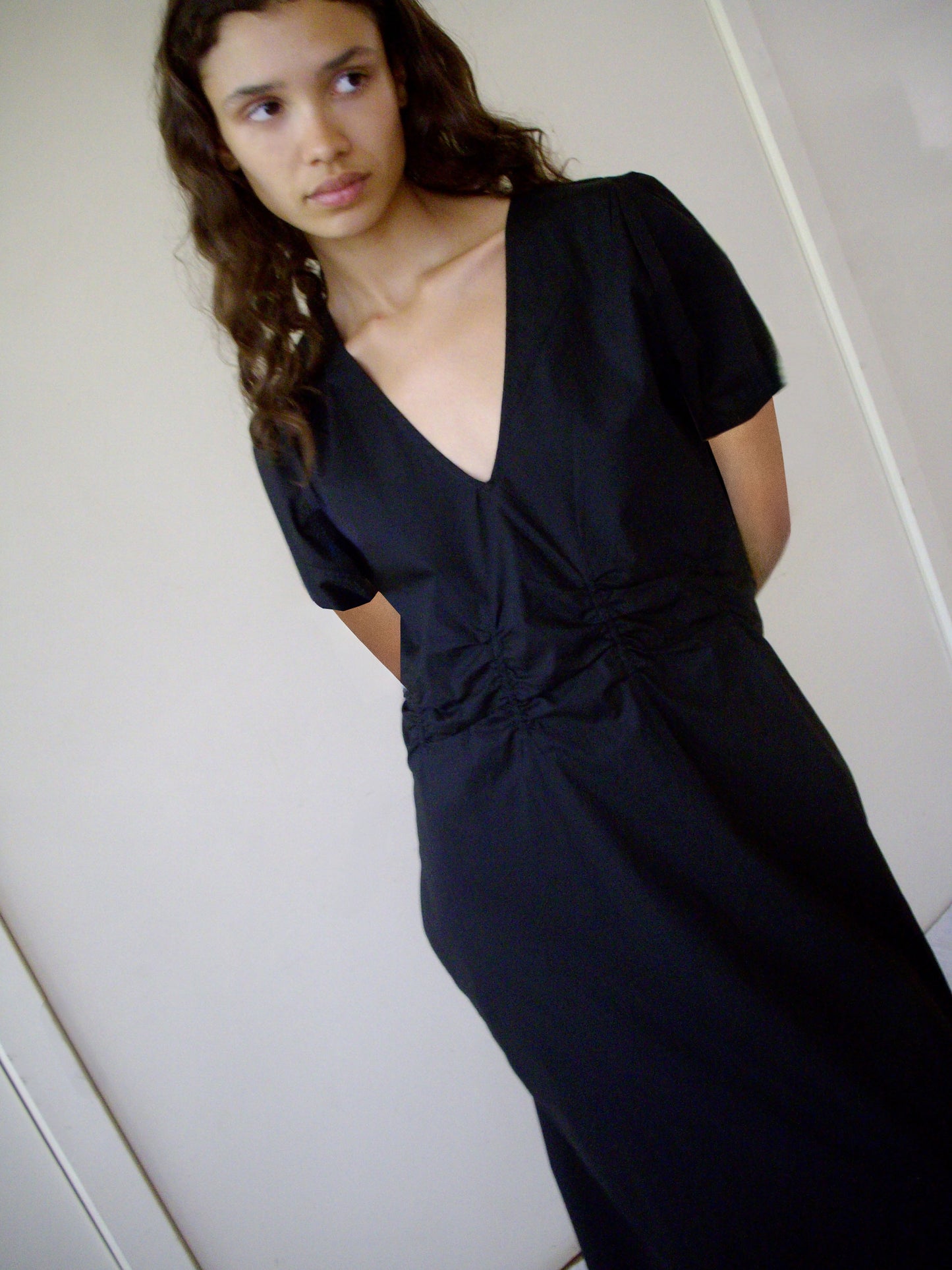 A young woman with long, wavy hair stands indoors with her hands behind her back, gazing slightly to the side while wearing the Pinched Dress - Black, made from organic cotton and featuring short sleeves and a deep V neckline.