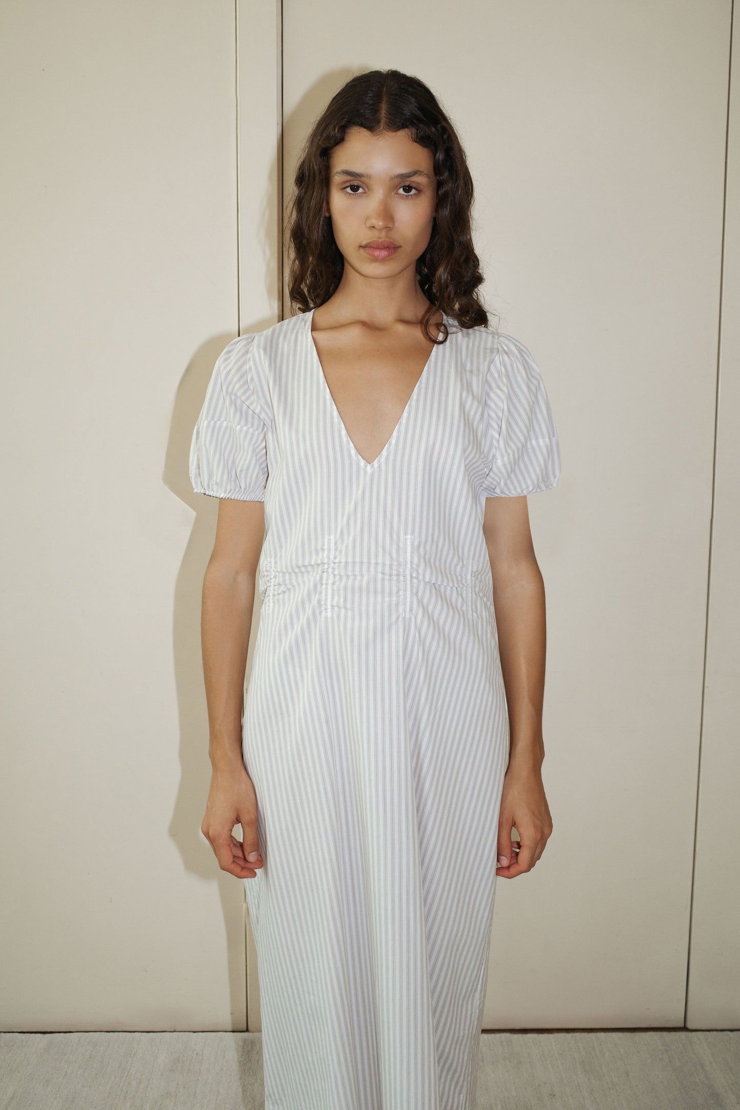 A woman with long, wavy brown hair wears the Pinched Dress - Lake Stripe, a light gray and white striped organic cotton poplin dress with short puff sleeves and a deep V-neckline, standing against a plain beige wall.
