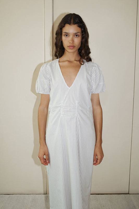A woman with long wavy hair stands against a beige wall, facing forward in the Pinched Dress—a white and light gray striped dress featuring short puff sleeves and a deep V-neckline, styled with a neutral expression.