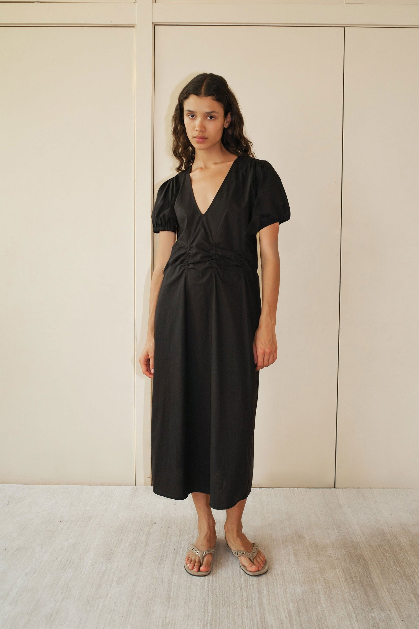 A woman with wavy, shoulder-length hair poses indoors in front of light paneled doors, wearing the Pinched Dress - Black—an organic cotton, short-sleeved midi dress with a V neckline—paired with beige sandals.