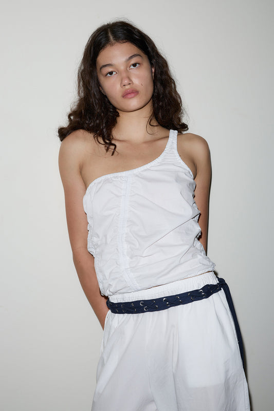 A young woman with wavy brown hair wears a white Scrunchie Top and loose white pants with a dark belt, standing against a plain light-colored wall, looking at the camera with a neutral expression.