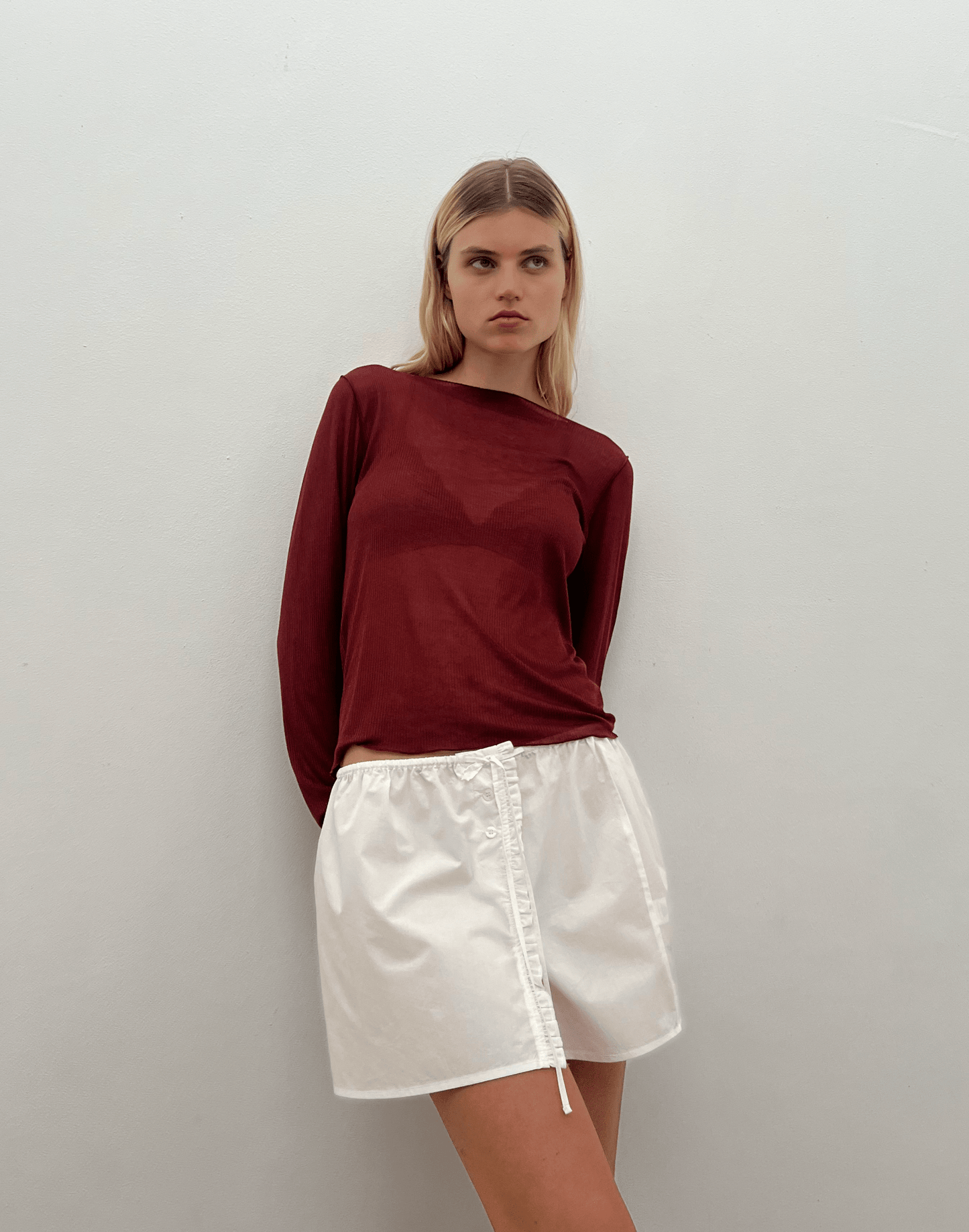 A woman with straight blonde hair and a neutral expression stands against a plain white wall, wearing the 0 Ruffle Mini Skirt in white and a long-sleeve burgundy top, with her arms behind her back.