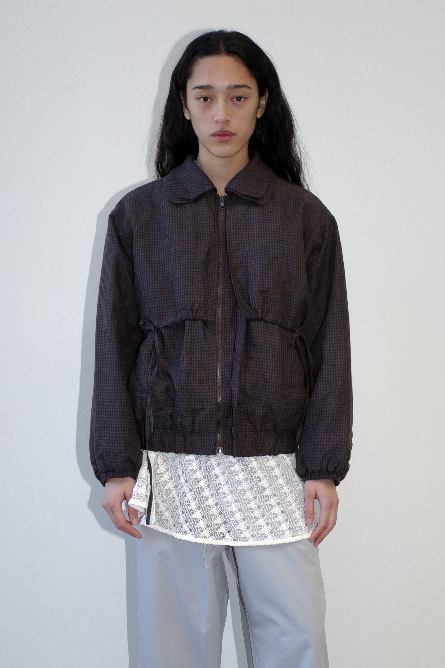 A person with long dark hair stands against a plain light wall, wearing the Double Layered Jacket - Slate Check over a white patterned shirt and light gray pants, looking directly at the camera with a neutral expression.