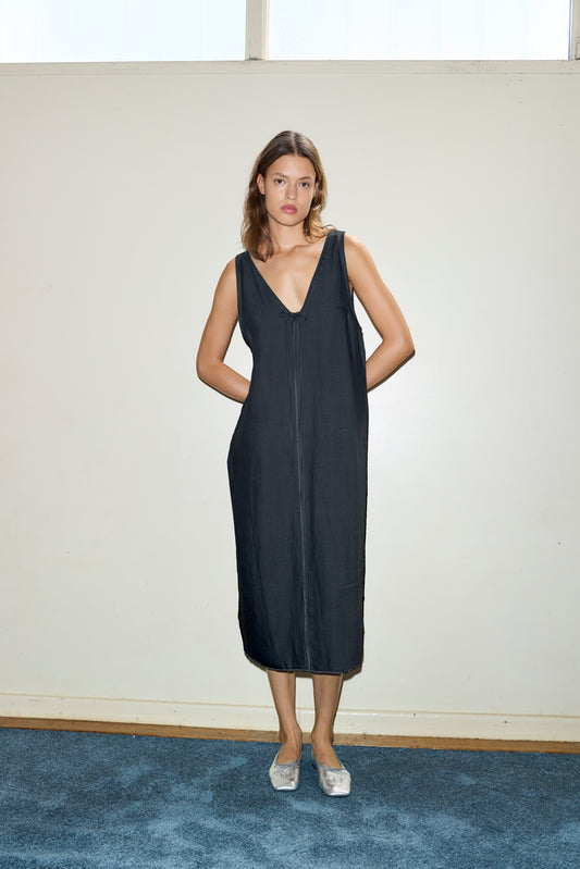 A woman stands indoors against a plain light-colored wall, barefoot on a blue carpet, wearing the Tie Slip Dress—a sleeveless, loose-fitting, deep V-neck black dress with pockets.