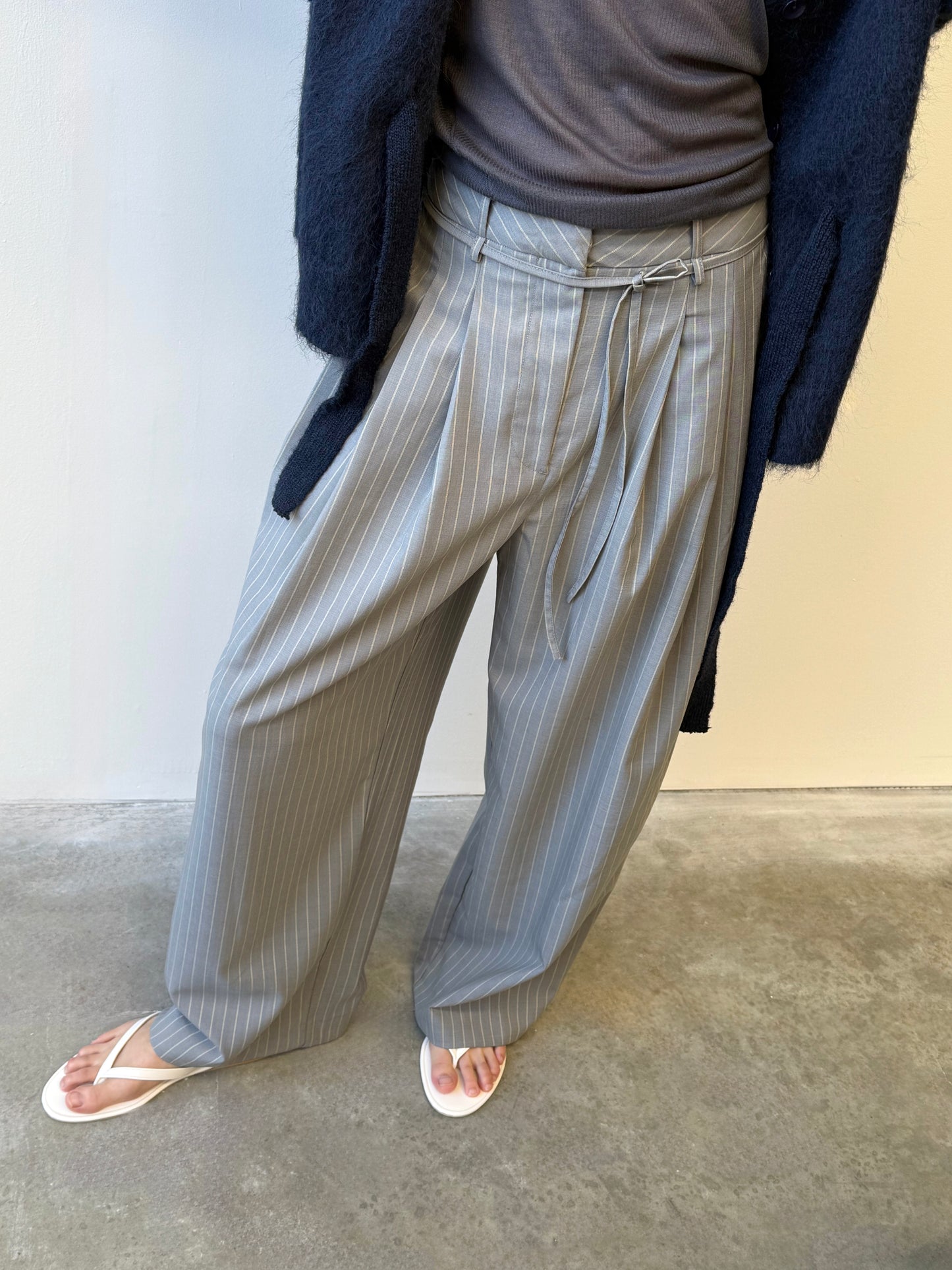 A person wearing the Tri Pleat Pure Wool Pant - Steele Stripe with soft front pleats, a dark top, navy cardigan, and white flip-flops stands on a concrete floor against a light-colored wall.