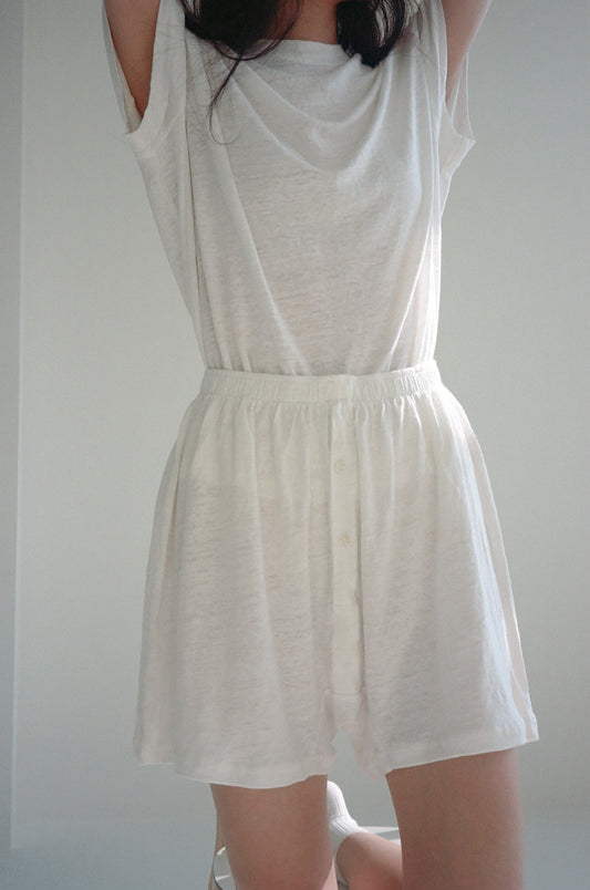 Model wears the soft short - ecru: high-waisted, linen boxer-style shorts paired with a loose, short-sleeved white top. She stands against a light background with arms raised and face out of frame.