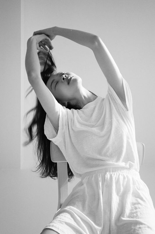 A woman in a Soft T-Shirt and shorts sits on a chair, eyes closed, arching her back with arms stretched overhead and hair cascading down. The black and white image highlights the comfort of the Soft T-Shirt.