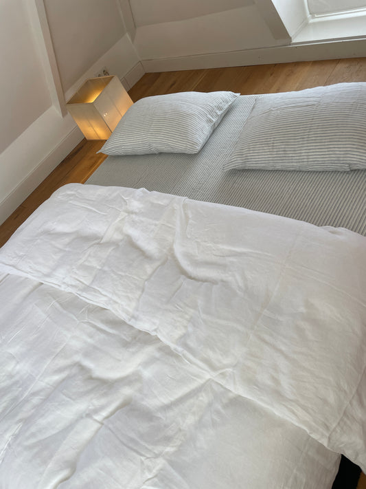 A neatly made bed with white sheets, two pillows, and the Duvet cover - White US sits near a corner with wooden flooring. By the window, a small square lamp glows on the floor beside the bed.