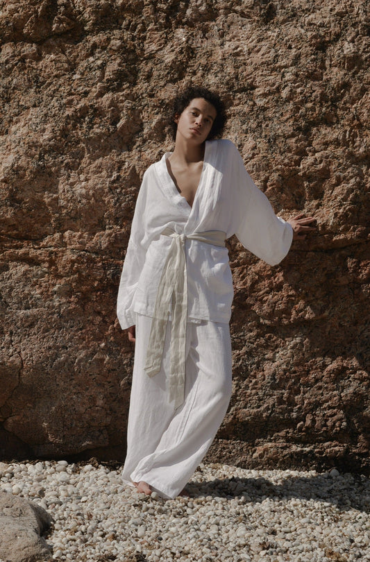 Someone wearing the 01 set - white, a loose organic linen loungewear outfit, stands on rocky ground against a rough stone wall, with natural sunlight illuminating the scene.