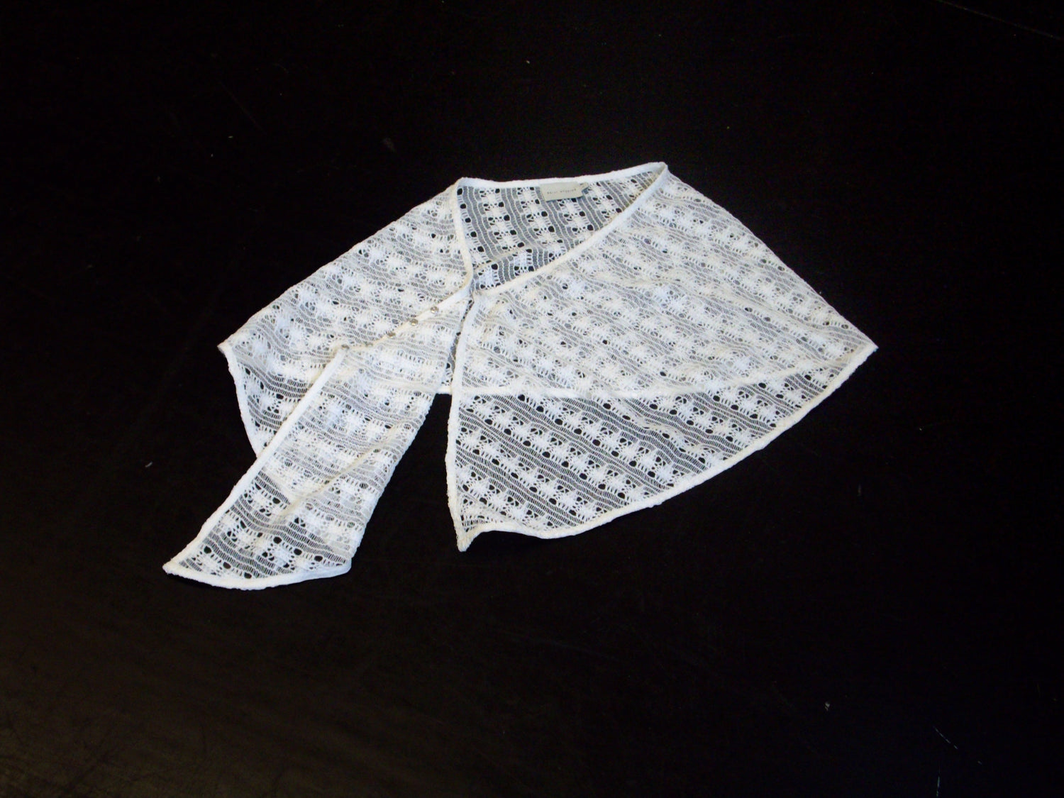 A white, lacy knitted shawl with a geometric openwork pattern is laid out on a solid black surface.