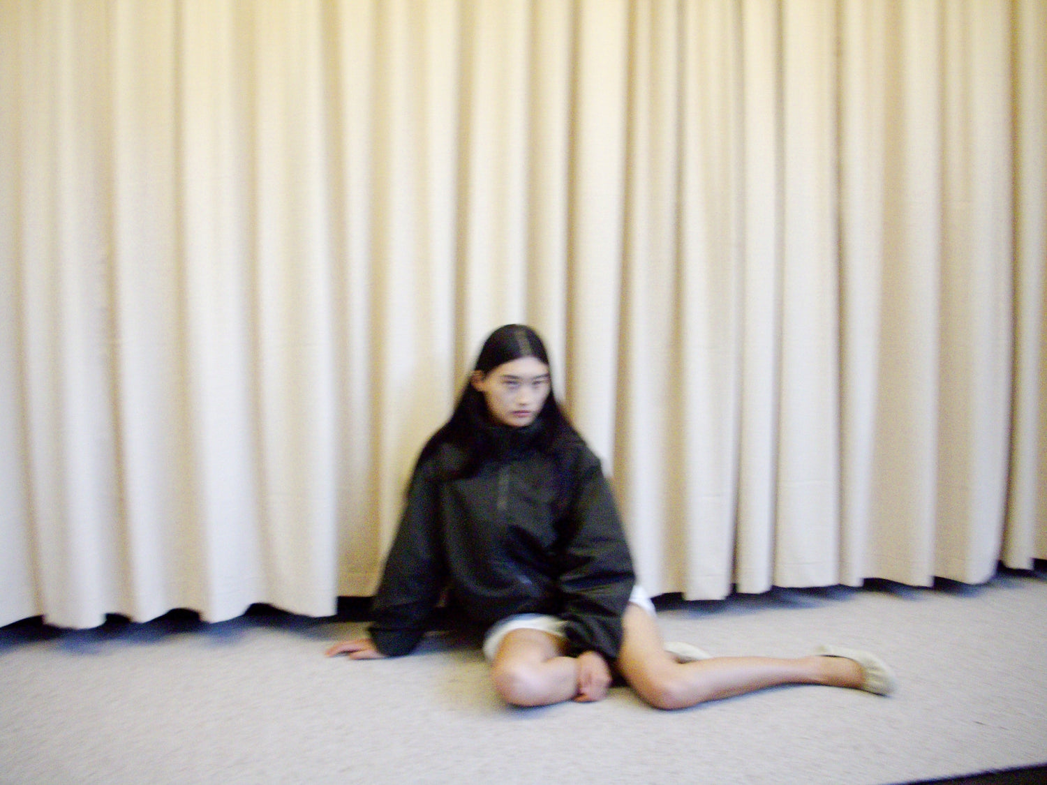 A person with long dark hair sits on the floor in front of beige curtains, wearing a dark oversized jacket and neutral shoes, looking off to the side.