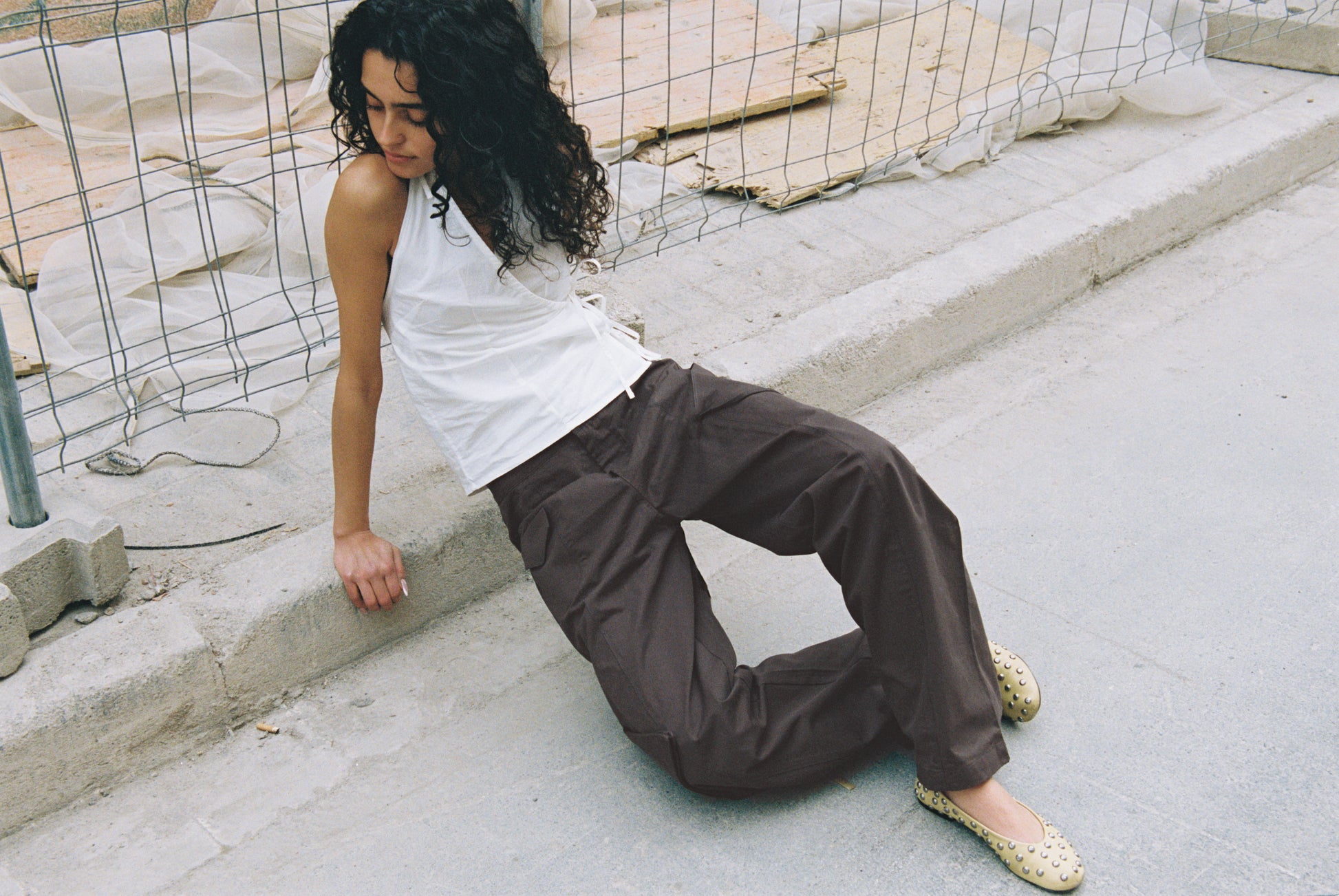 A woman with curly hair, wearing a white sleeveless top and the Pocket Pant - Wood made from organic cotton twill, sits by a wire fence at a construction site, looking down in a relaxed pose.
