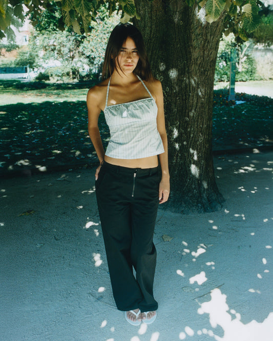 A woman stands under a leafy park tree, wearing a light-colored halter top and the Zip Pant - Black made from organic cotton twill. Dappled sunlight highlights her outfit, casting soft shadows on the ground.