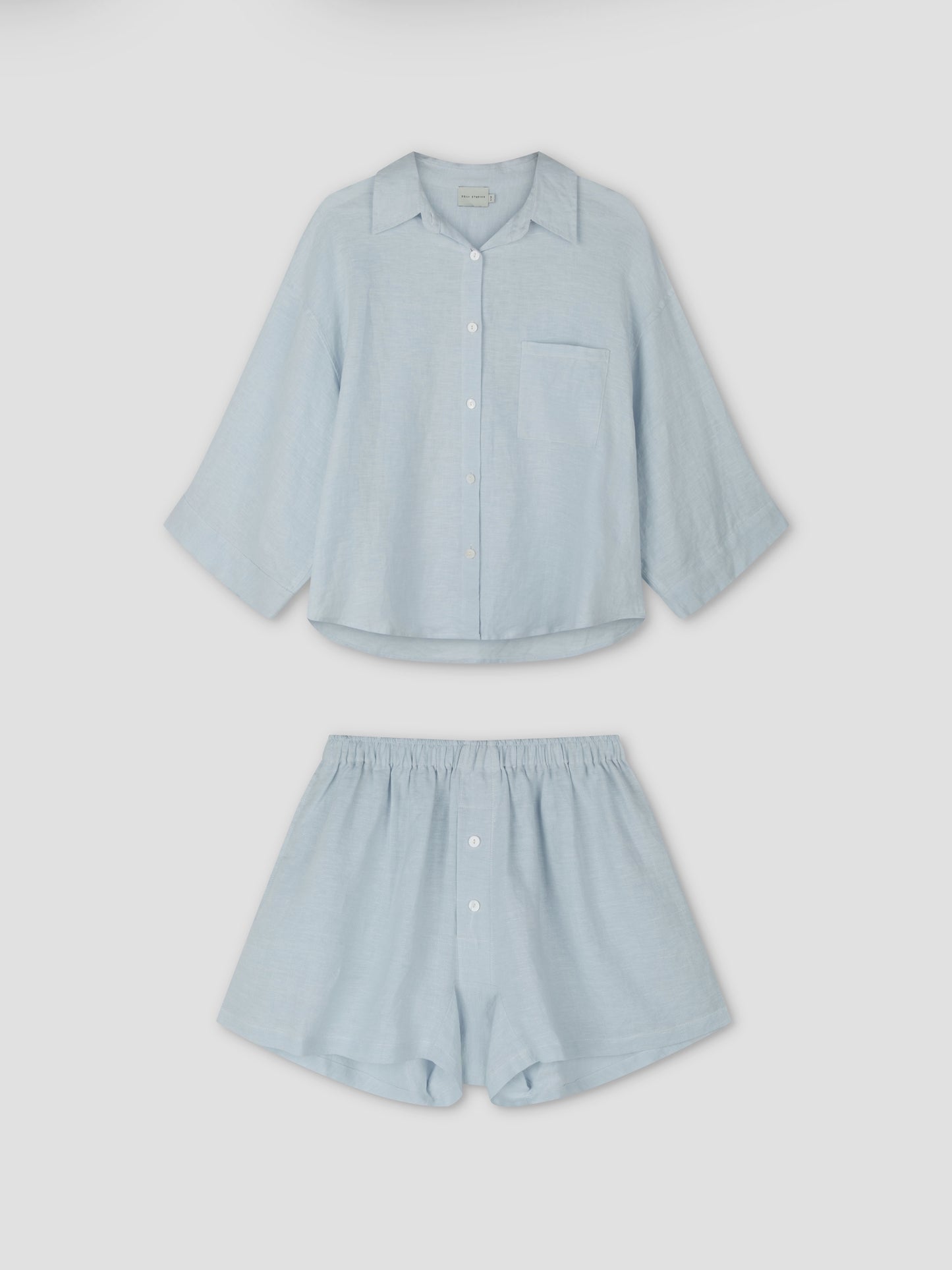The 03 Set - Soft Blue by Deiji Studios features an oversized light blue linen shirt with a chest pocket and matching boxer-style shorts with front buttons, shown on a plain white background.