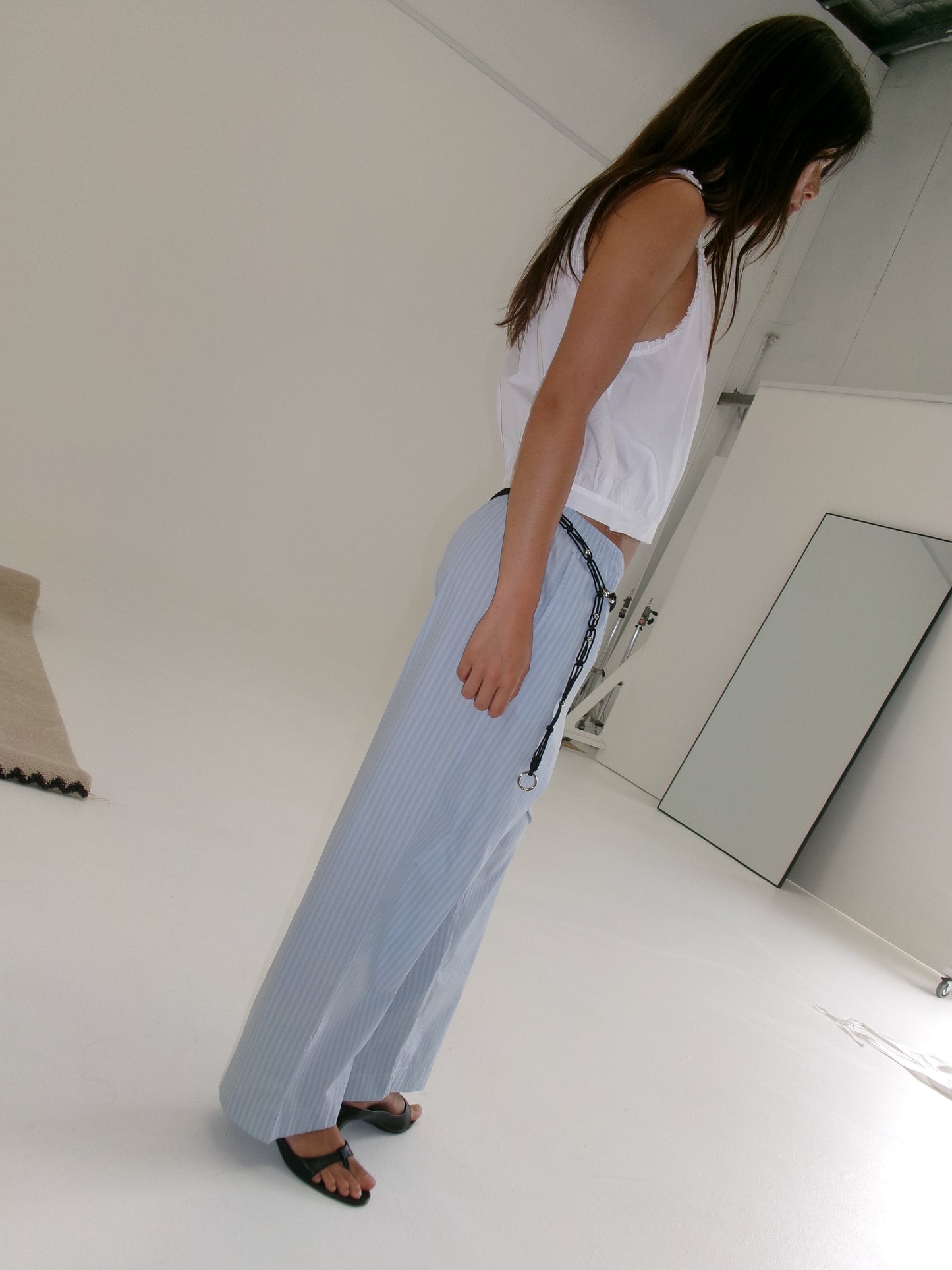 A woman with long brown hair wears the Ease Trouser - Lake Blue Stripe by Deiji Studios, styled with a black cord belt, white sleeveless top, and black sandals in a minimal white room as she faces right.