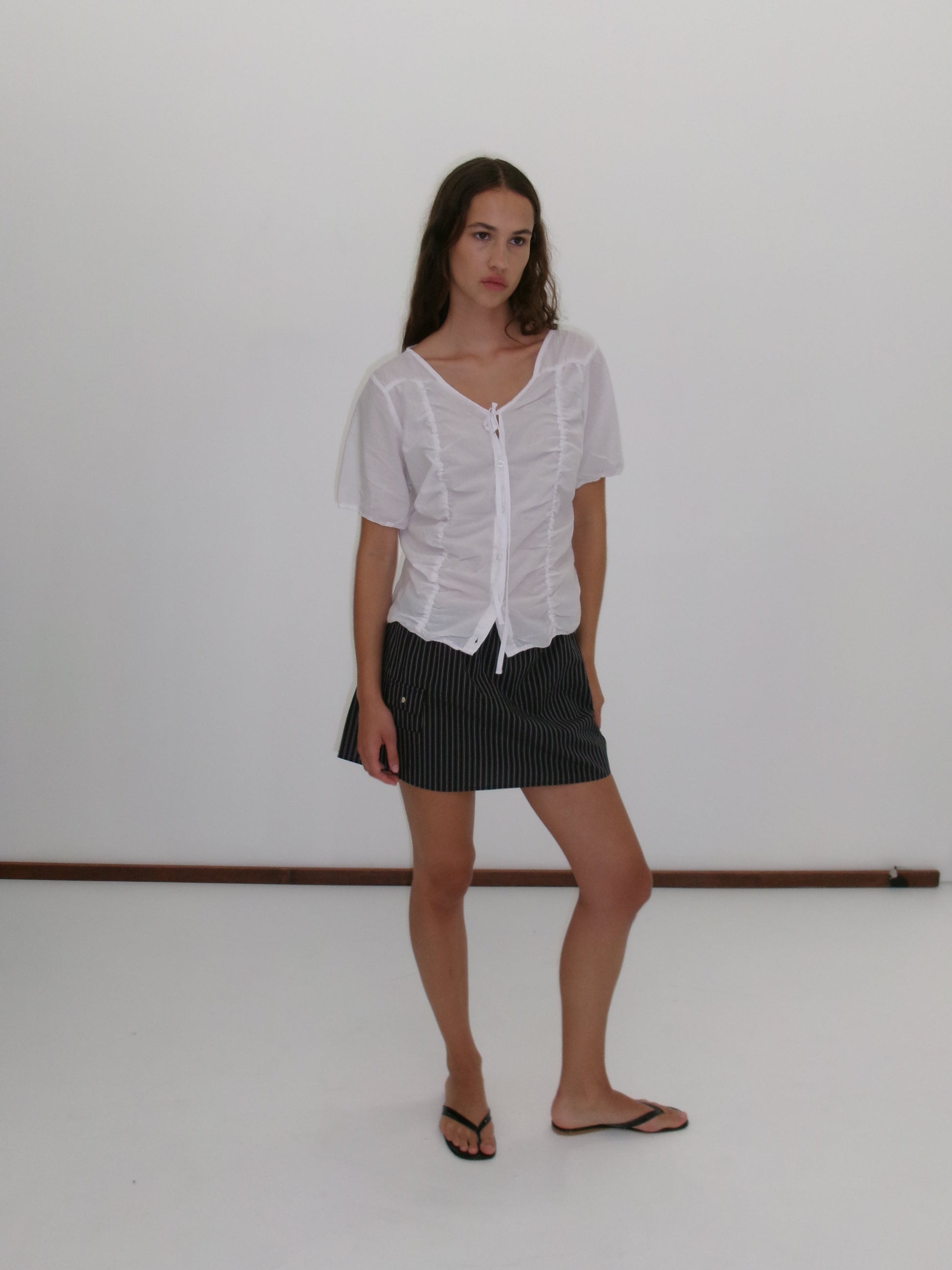 A woman with long brown hair and a neutral expression stands against a plain white background, wearing the Crinkle Top - White by Deiji Studios, a black pinstripe mini skirt, and black flip-flops.
